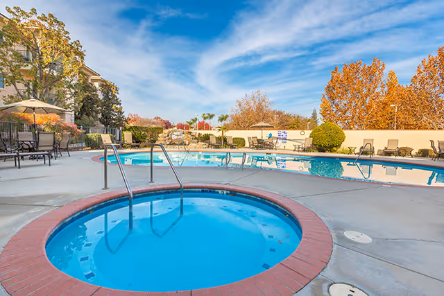 Outdoor pool area at a senior living facility with a circular hot tub in the foreground and a larger swimming pool in the background. There are lounge chairs and tables with umbrellas around the pool. Trees with autumn foliage and a clear blue sky are visible.