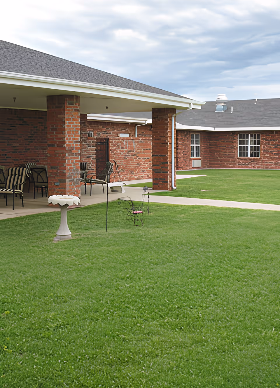 Outdoor courtyard area with green grass, a birdbath, metal chairs, and a covered patio supported by brick columns. The surrounding building has red brick walls and multiple windows under a cloudy sky.