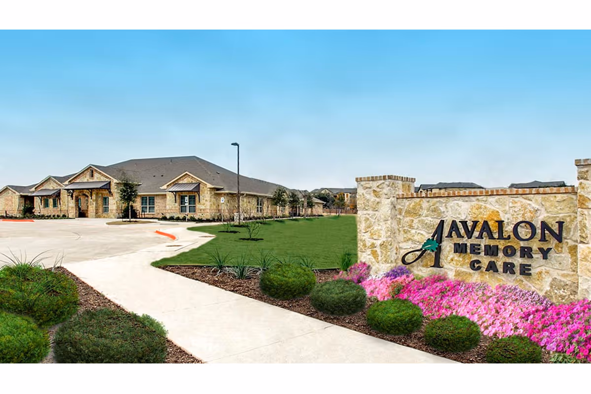 Exterior view of Avalon Memory Care facility showing a stone sign with the facility name, a landscaped garden with green bushes and pink flowers, a paved walkway, and a single-story building with a gray roof under a clear blue sky.