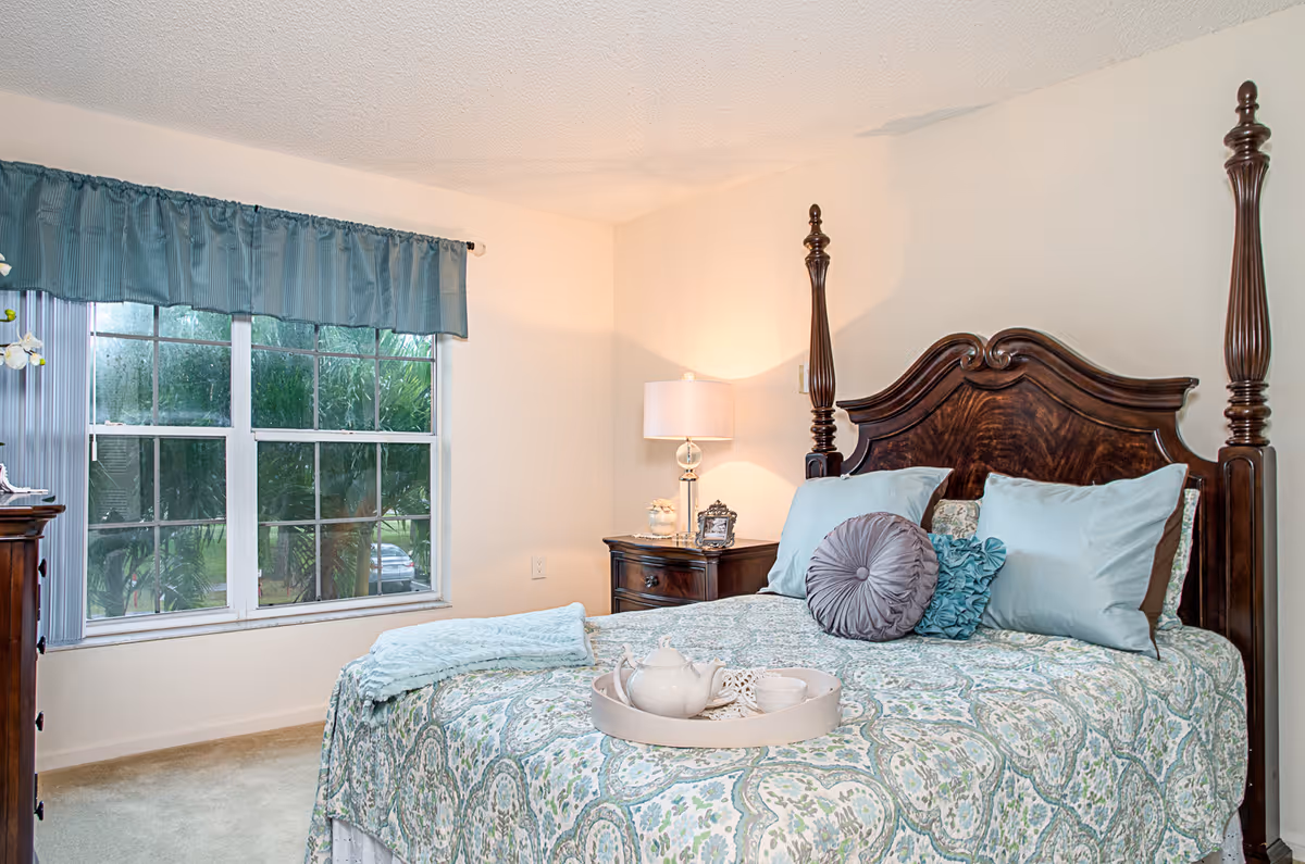 A cozy bedroom with a large wooden four-poster bed adorned with light blue and patterned bedding, decorative pillows, and a tray with a teapot and cups. There is a wooden nightstand with a lamp and decorative items beside the bed. A large window with blue curtains allows natural light to enter the room.