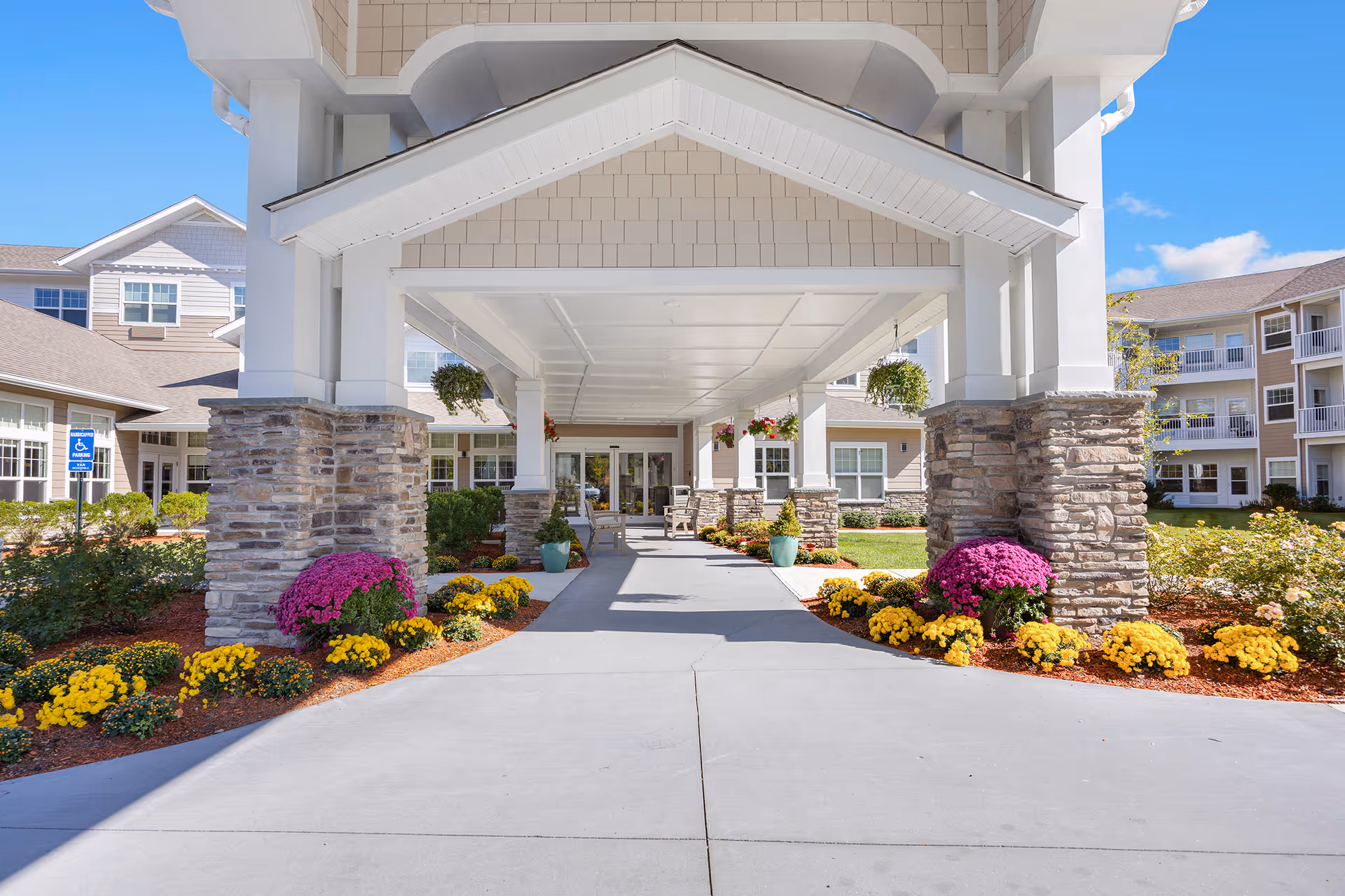 Covered entrance/porte-cochere of a senior living facility with stone columns, hanging plants, and colorful flowerbeds leading to glass doors.