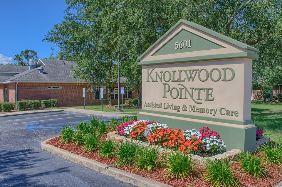 Outdoor view of the entrance sign for Knollwood Pointe Assisted Living and Memory Care, surrounded by flowers and greenery, with a brick building and parking spaces in the background under a clear blue sky.