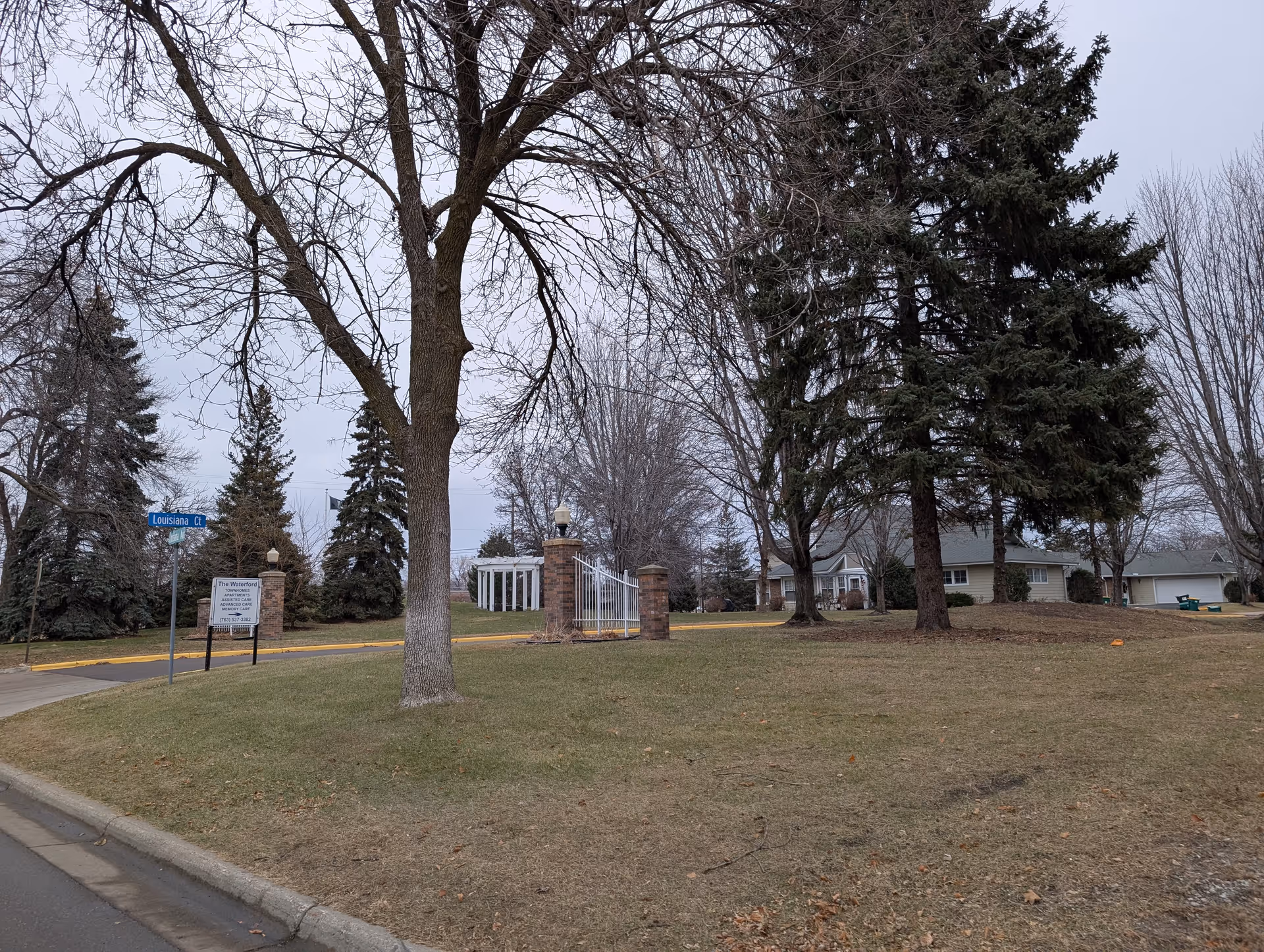 Outdoor view of Waterford Manor facility showing a grassy area with leafless trees and evergreen trees. There is a brick and white metal gate entrance, a street sign for Louisiana Ct, and a sign for The Waterford Apartments, Assisted Care, and Memory Care. Residential buildings are visible in the background under an overcast sky.
