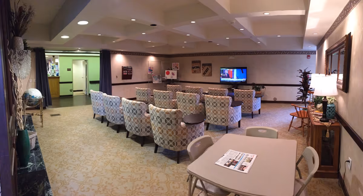 A cozy common room with patterned armchairs arranged in rows facing a wall-mounted television. There is a table with chairs in the foreground with a newspaper on it. The room has beige walls, carpeted floor with a subtle pattern, and decorative elements including a large mirror, lamps, plants, and framed pictures on the walls.