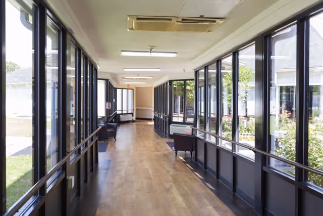 Sunlit interior corridor with large windows, handrails, wood flooring, and a few chairs overlooking a garden.