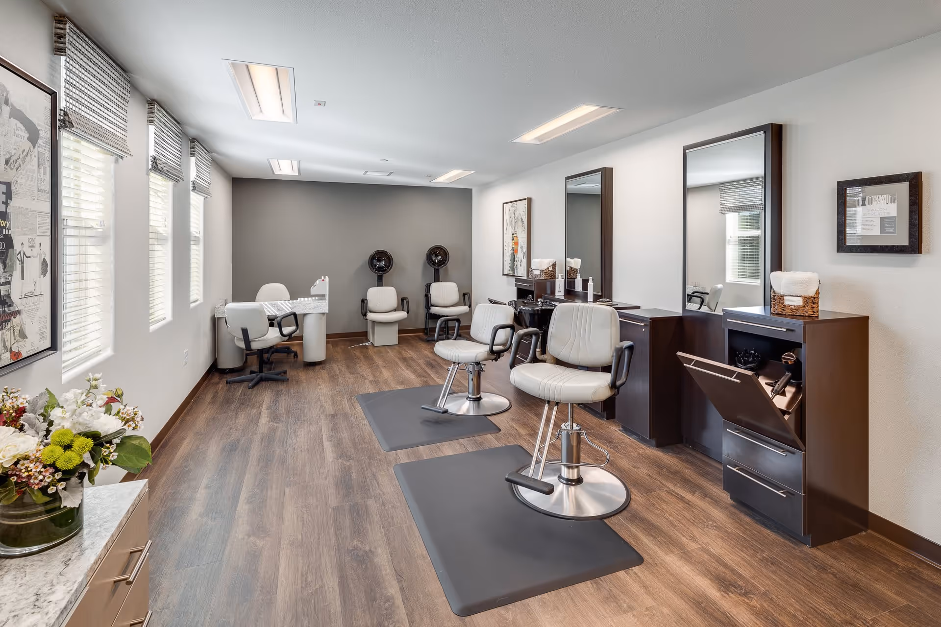 Interior view of a modern salon room with two styling chairs on black mats in front of large mirrors, two hair dryer chairs against a gray accent wall, a manicure table with chairs, and a cabinet with a hair dryer. The room has wood flooring, white walls, and several windows with blinds letting in natural light.