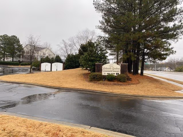 Entrance sign for Lakeside Gardens on a grassy, tree-lined median beside a wet road on an overcast day.