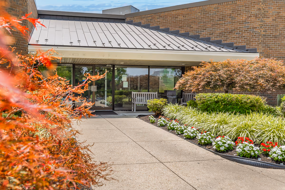 Entrance to Autumn Lake Healthcare at Patuxent River with a concrete walkway leading to glass double doors. The entrance is surrounded by well-maintained landscaping including green shrubs, red and white flowers, and trees with orange leaves. The building exterior is made of brown brick with a metal roof over the entrance.