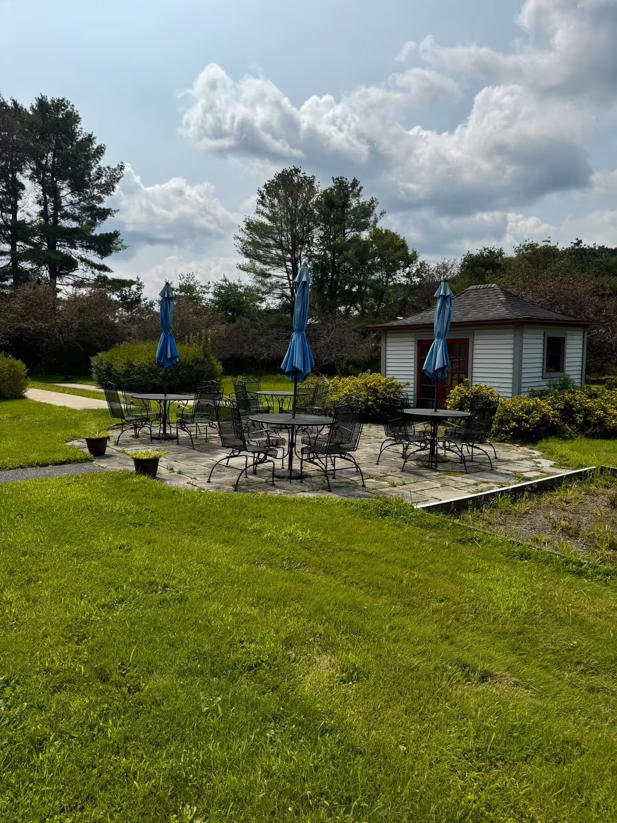 Outdoor patio area with several round metal tables and chairs, each table shaded by a closed blue umbrella. The patio is surrounded by green grass and bushes, with a small white building with a dark roof in the background. Trees and a partly cloudy sky are visible behind the scene.