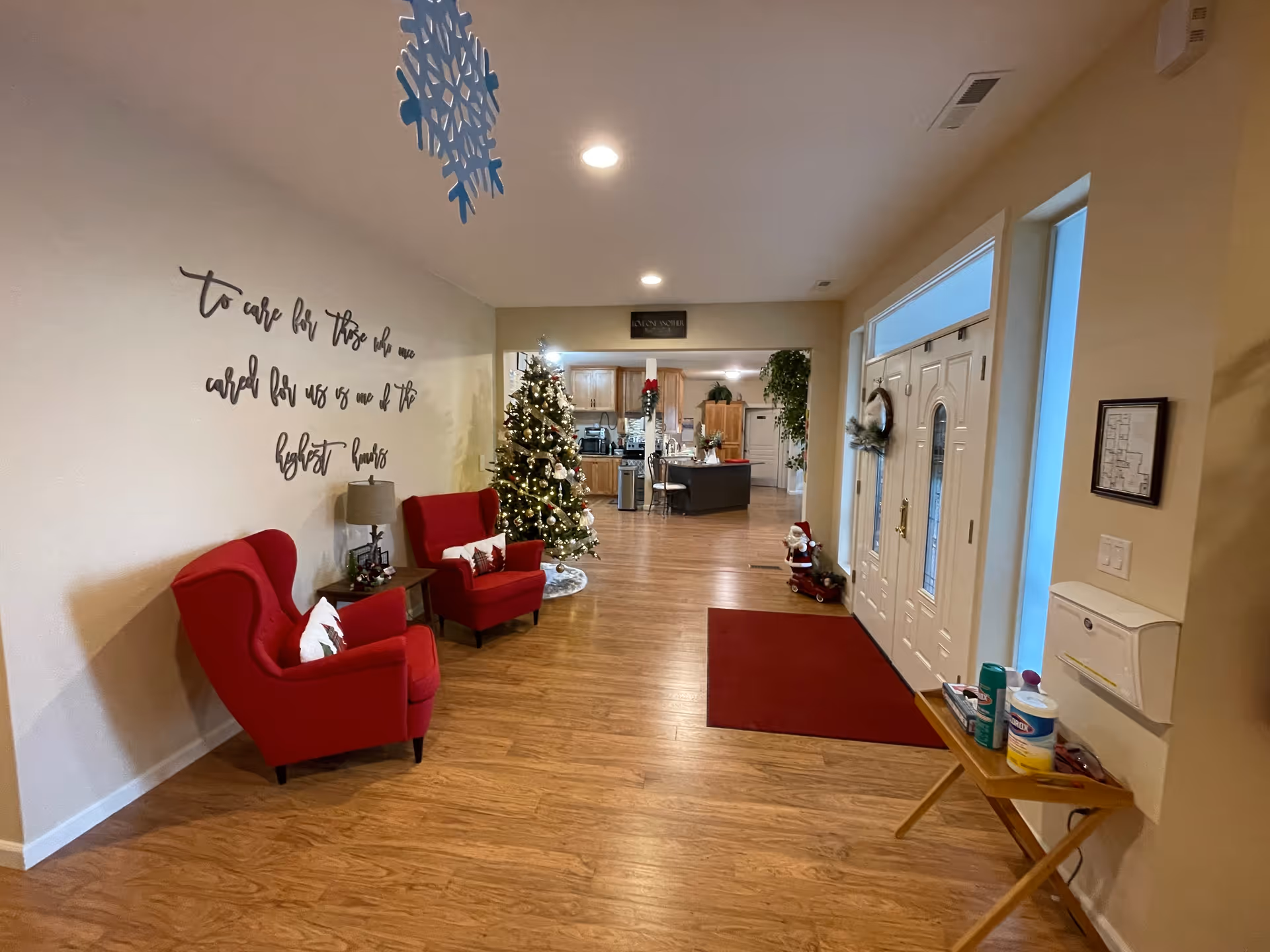 Interior view of a senior living facility entrance area decorated for Christmas. Two red armchairs with white pillows are placed against a wall with a decorative quote. A decorated Christmas tree stands near the chairs. The entrance door is white with glass panels and a wreath. A small table near the door holds cleaning supplies. The floor is wooden, and the space opens into a kitchen area in the background.