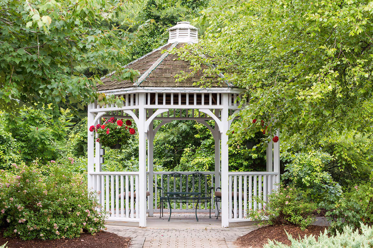 A white wooden gazebo with a shingled roof situated in a lush garden surrounded by green trees and bushes. There are hanging flower baskets with red flowers on the gazebo, and a black metal bench inside it on a paved stone pathway.