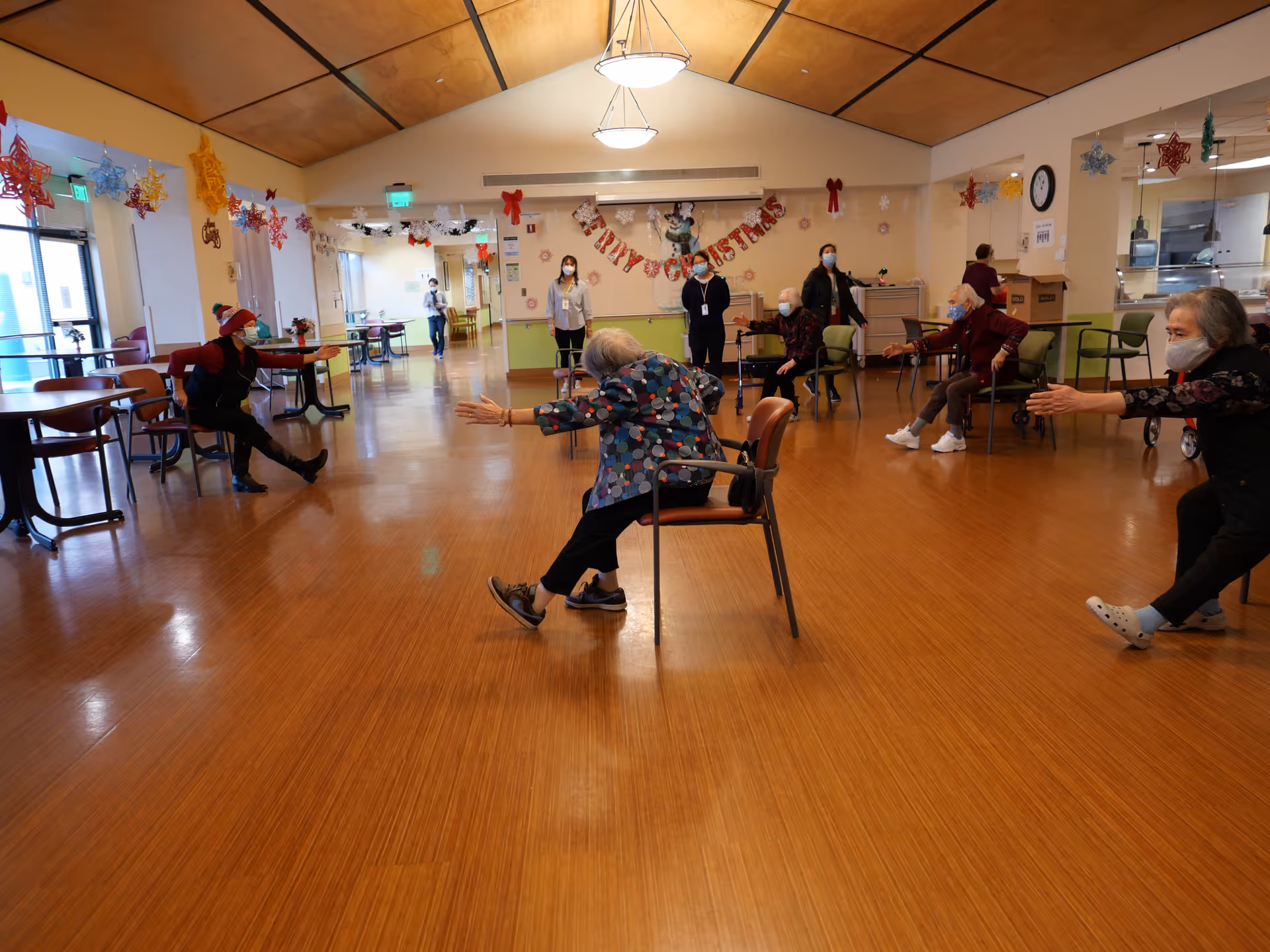 A group of elderly people wearing masks participating in a seated exercise class in a spacious, well-lit room decorated with Christmas ornaments and a 'Merry Christmas' banner. Some participants are seated on chairs while others are standing, stretching their arms and legs. Staff members are present in the background.