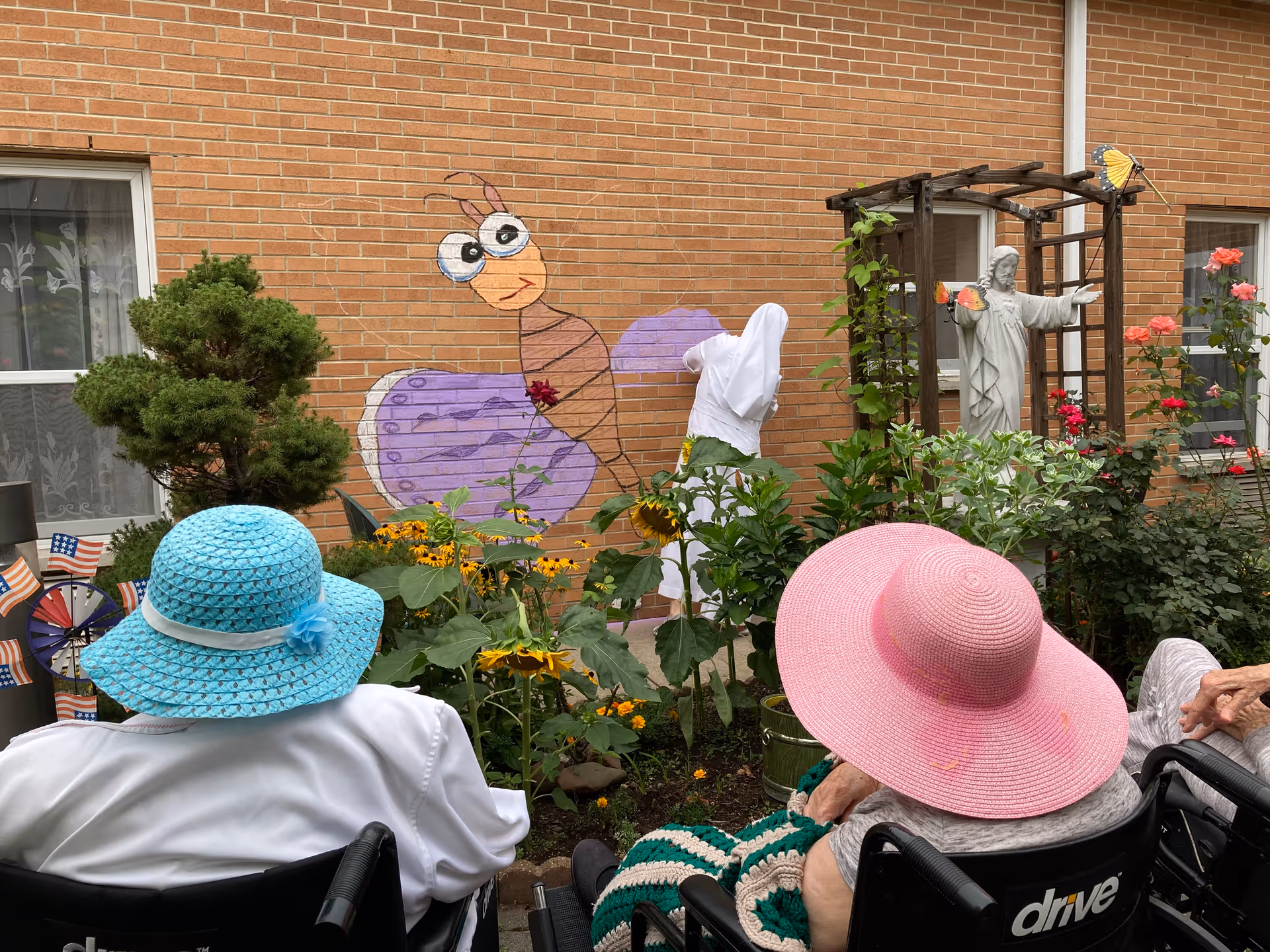 Two elderly individuals in wheelchairs wearing large sun hats, one blue and one pink, are sitting in a garden area with various flowers and plants. A brick wall with a colorful cartoon butterfly mural and a statue of Jesus with butterflies on it is visible in the background.