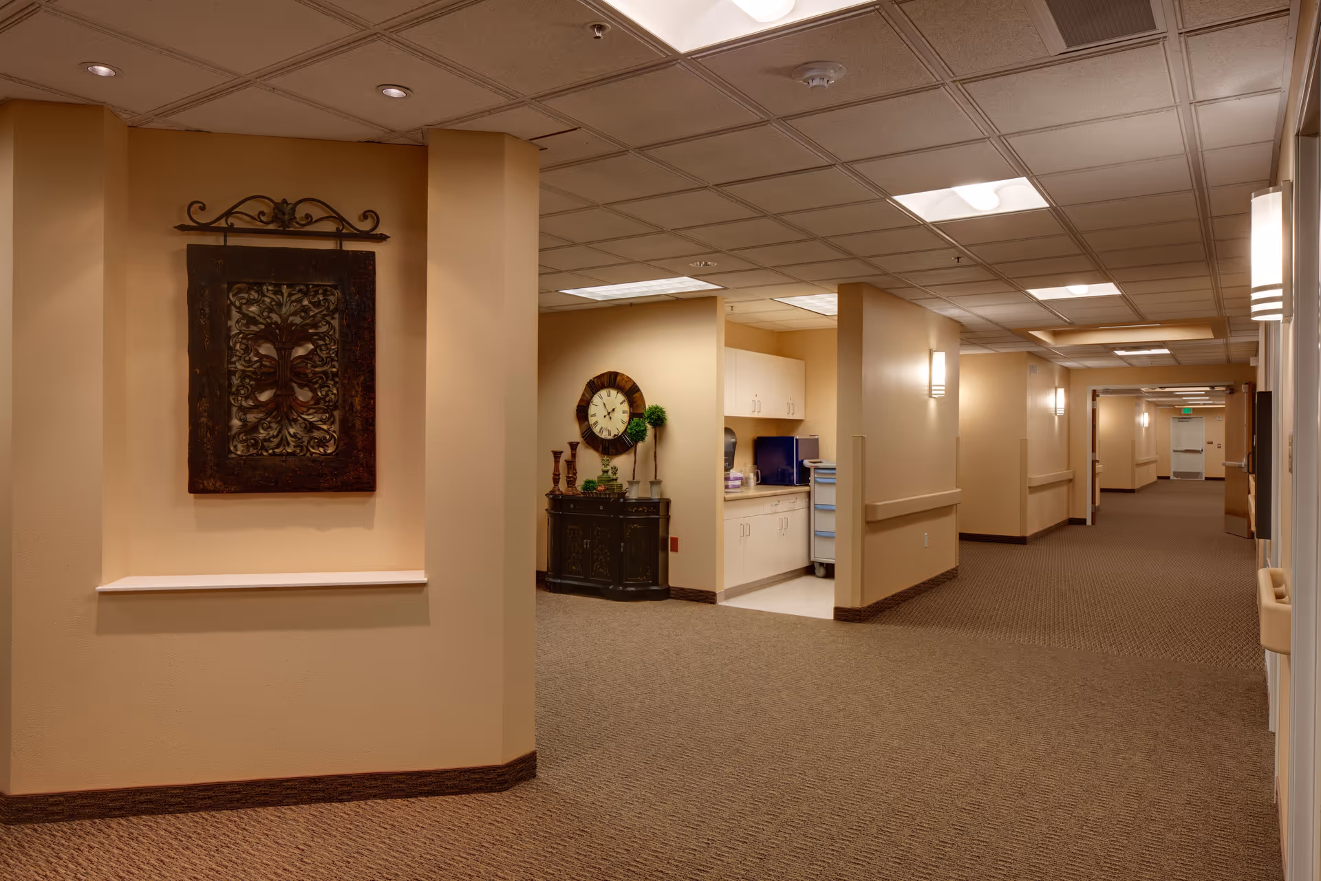 Carpeted interior hallway with recessed lighting, wall sconces, decorative wall art, a small cabinet with a clock and cabinets set into a nursing station area.