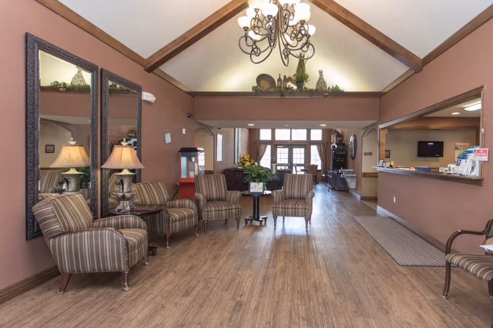 A spacious senior living facility common area with wooden flooring, striped armchairs arranged around small tables, two large mirrors on the left wall, a chandelier hanging from a vaulted ceiling with wooden beams, and a reception desk on the right side. The area is well-lit with natural light coming from large windows in the background.