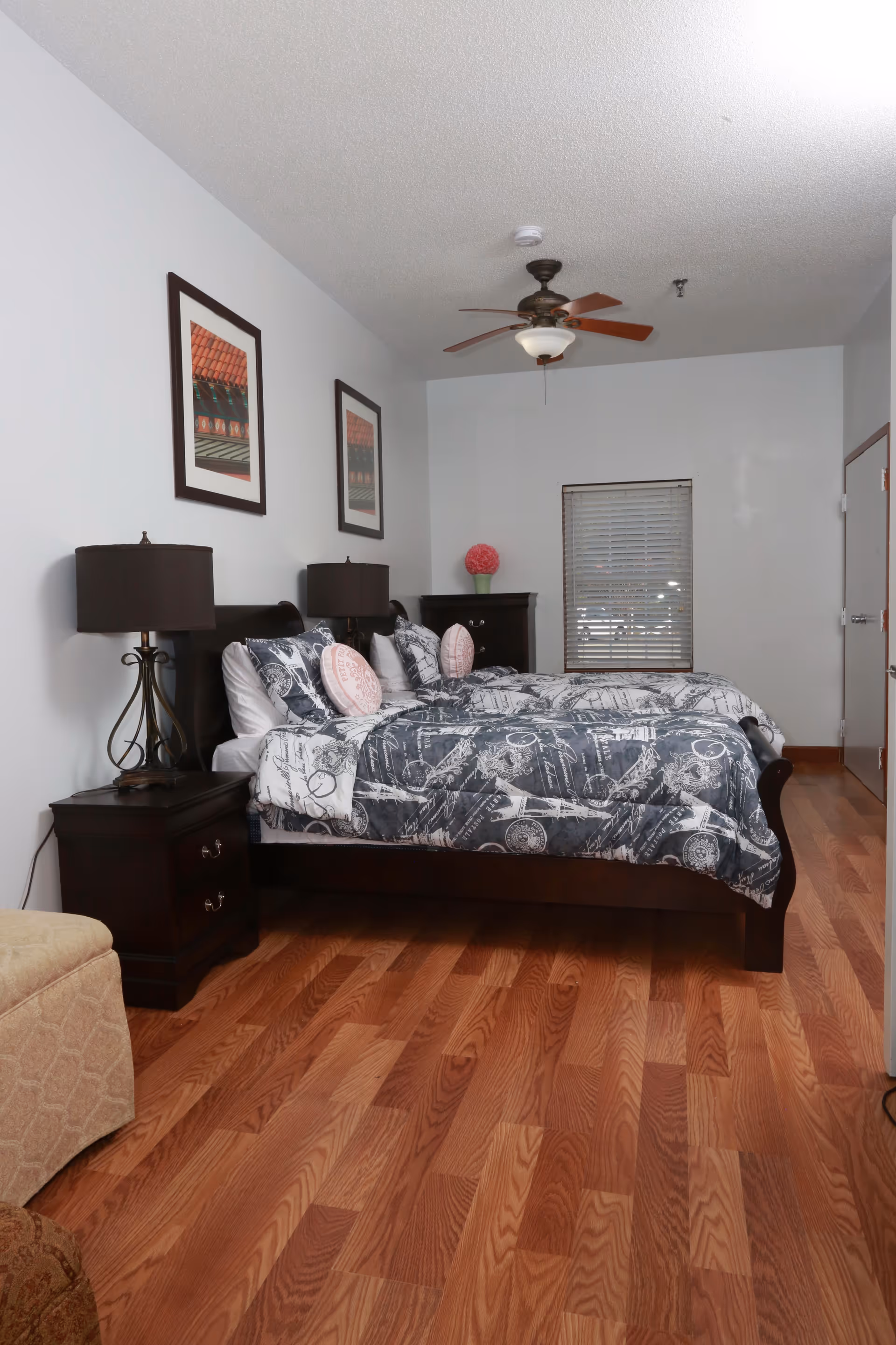 A bedroom with two twin beds featuring dark wooden frames and matching nightstands with lamps. The beds have patterned bedding in shades of gray and white with decorative pillows. The room has wooden flooring, a ceiling fan with light, two framed pictures on the wall, a window with blinds, and a dark wooden dresser with a pink flower arrangement on top.