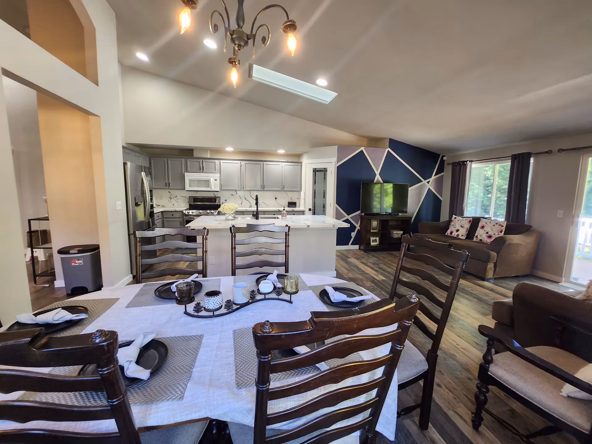 Interior view of a senior care home showing a dining area with a table set with placemats, plates, and napkins, a kitchen with gray cabinets, stainless steel appliances, and a white island, and a living room area with a brown sofa, a TV on a stand, and a wall painted with geometric patterns in blue and white. Large windows with curtains allow natural light into the space.