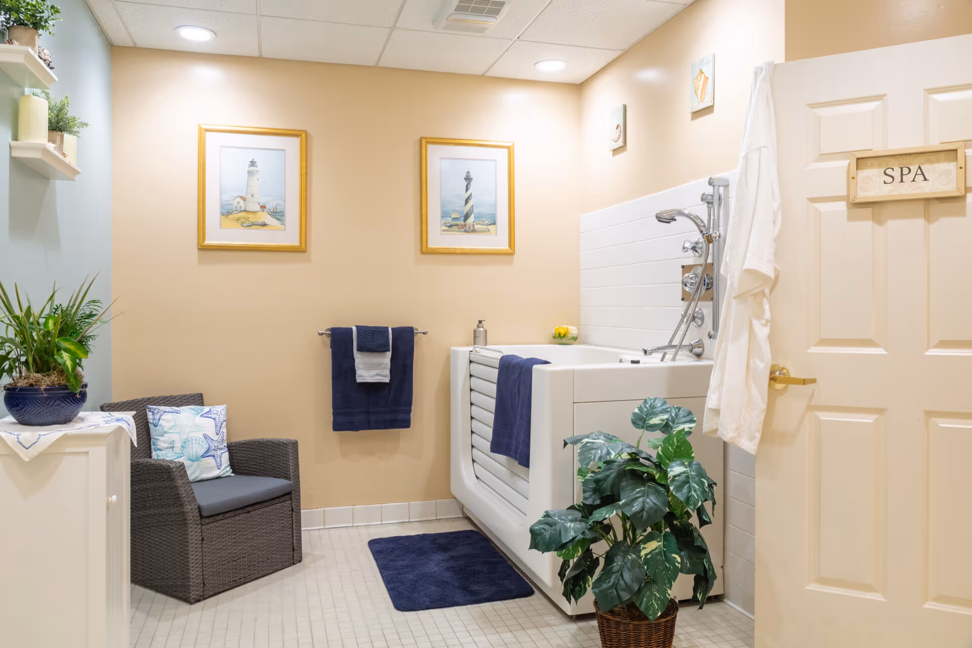 A spa room with a large white bathtub equipped with multiple shower heads and controls. The room has beige walls with two framed lighthouse pictures hanging above a towel rack holding navy blue towels. There is a wicker chair with a cushion and a pillow featuring starfish designs, a small cabinet with potted plants, and a green leafy plant in a basket on the floor. A white robe hangs on the door, which has a wooden sign labeled 'SPA'.