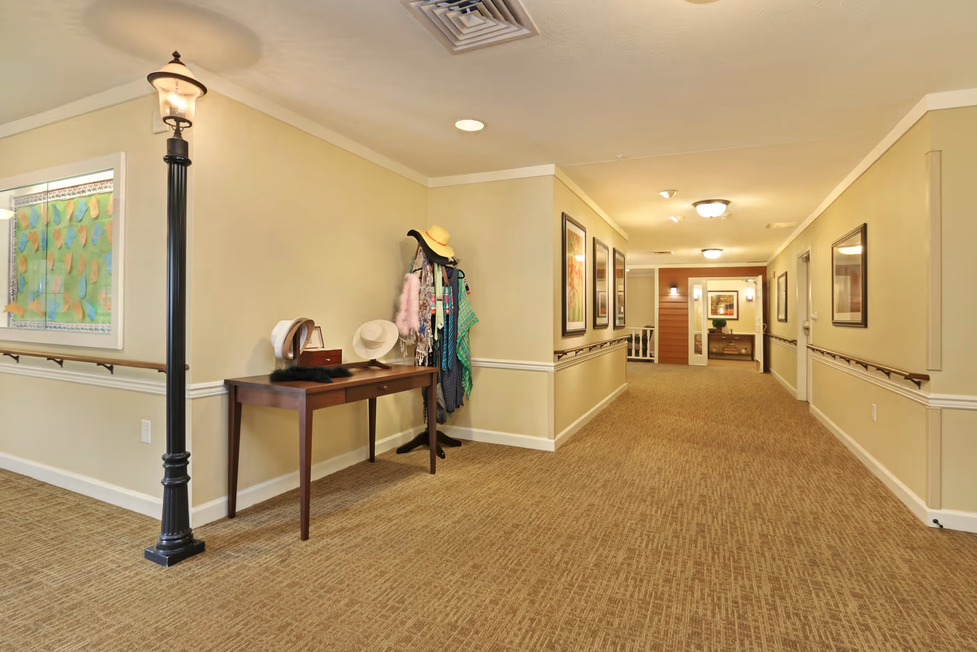 A well-lit hallway in a senior living facility with beige walls and carpeted floor. On the left side, there is a decorative black streetlamp and a wooden table with hats and scarves displayed. A coat rack with various hats and scarves stands next to the table. The hallway is decorated with framed artwork on the walls and has handrails on both sides.