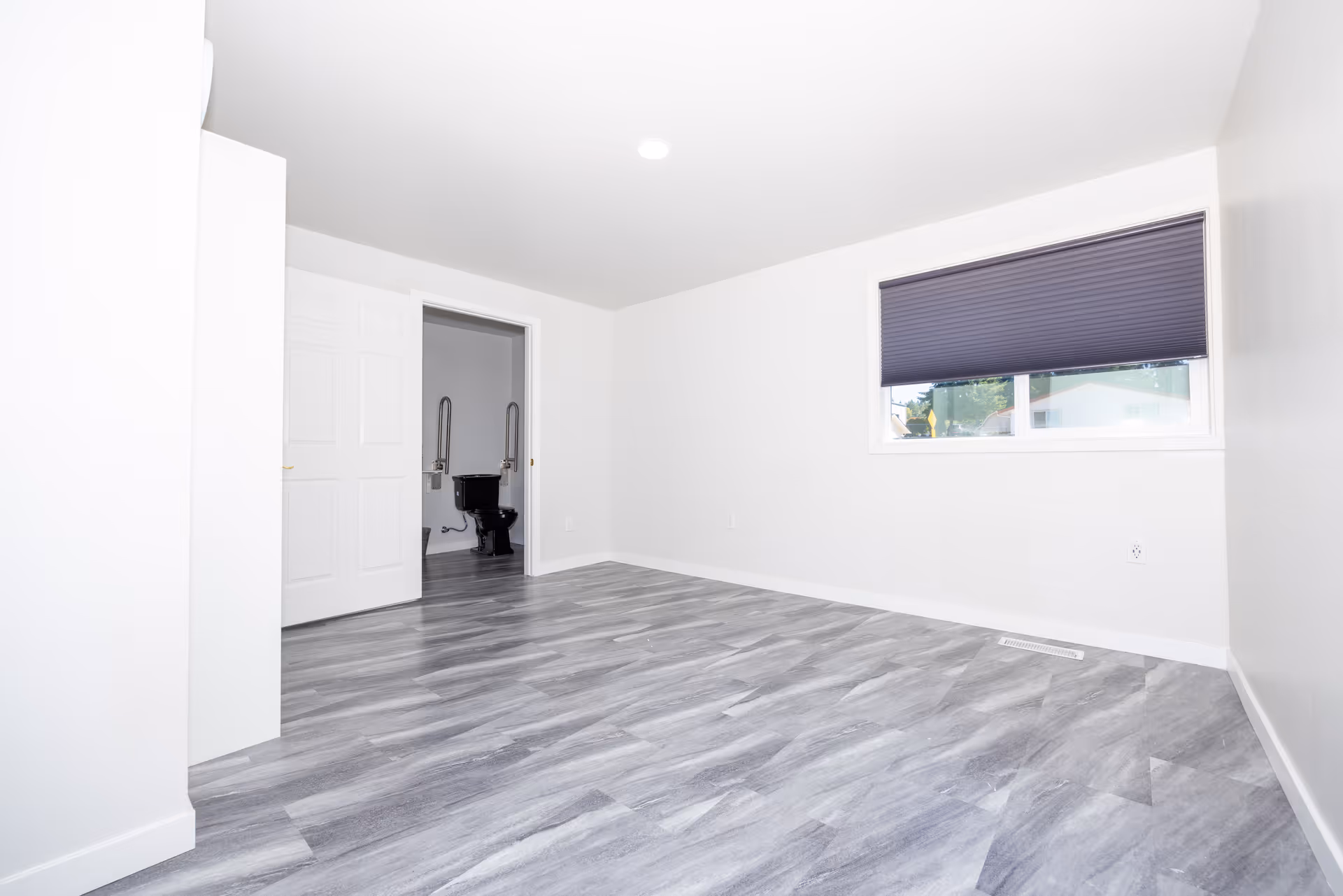 Empty room with gray wood-patterned flooring, white walls, a window with a dark blind, and an open door leading to a bathroom with a black toilet and grab bars.