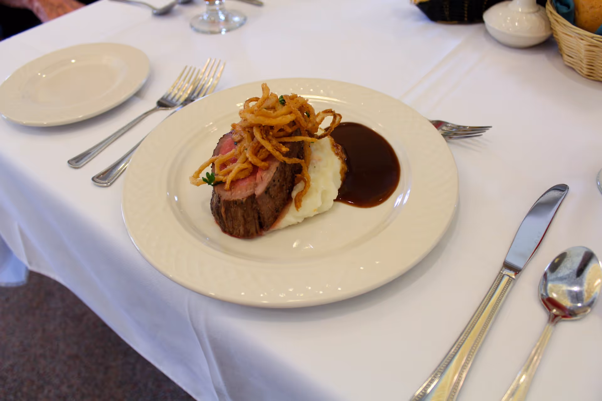 A plated meal on a white tablecloth featuring a slice of roast beef topped with crispy fried onions, served with mashed potatoes and brown gravy. The table is set with silverware, a bread basket, and a small white condiment container.