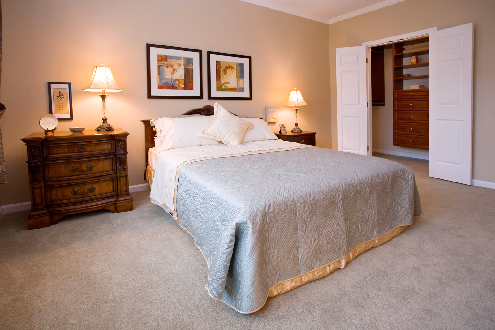 A neatly made bed with a light blue quilt and white pillows in a bedroom. There are two wooden nightstands on either side of the bed, each with a lamp. Above the bed are two framed floral artworks. To the right, there is an open closet with wooden shelves and drawers. The room has beige walls and carpeted floor.