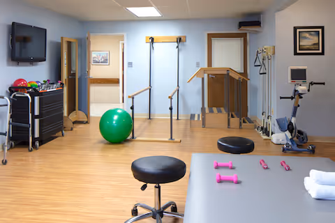 Well-lit therapy/fitness room with parallel bars, a treatment table, exercise ball, hand weights, and a stationary bike.