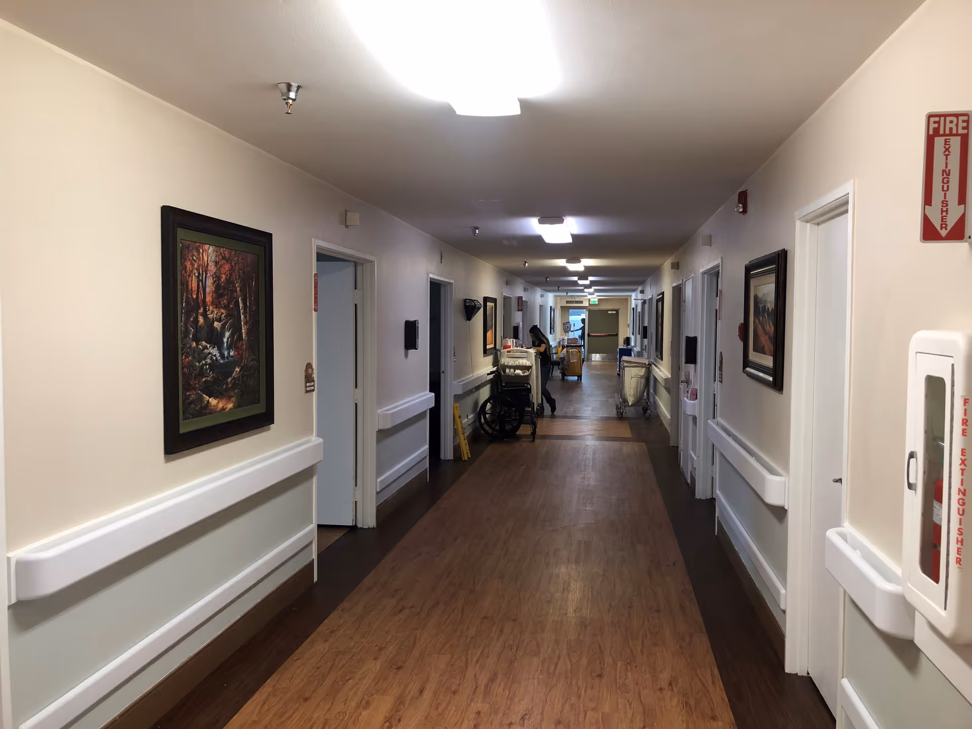 Long interior corridor of a senior living facility with wood-look flooring, wall handrails, framed artwork and medical carts at the far end.