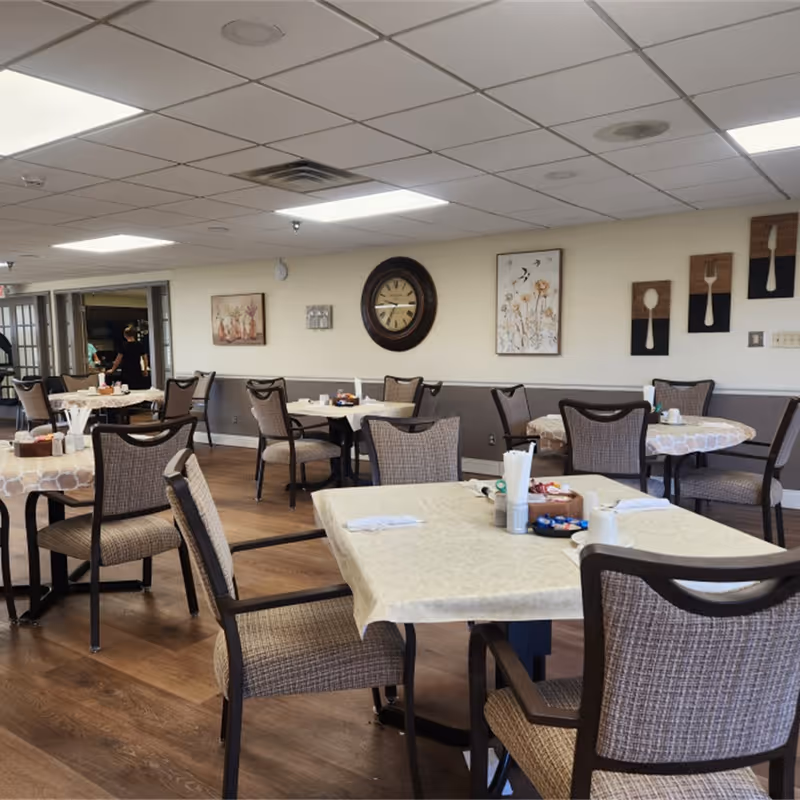 A dining room in an assisted living facility with several tables covered with tablecloths and surrounded by cushioned chairs. The walls are decorated with a large clock, floral artwork, and wooden panels featuring utensils. The floor is wooden, and the ceiling has recessed lighting. In the background, a person is seen near a doorway.