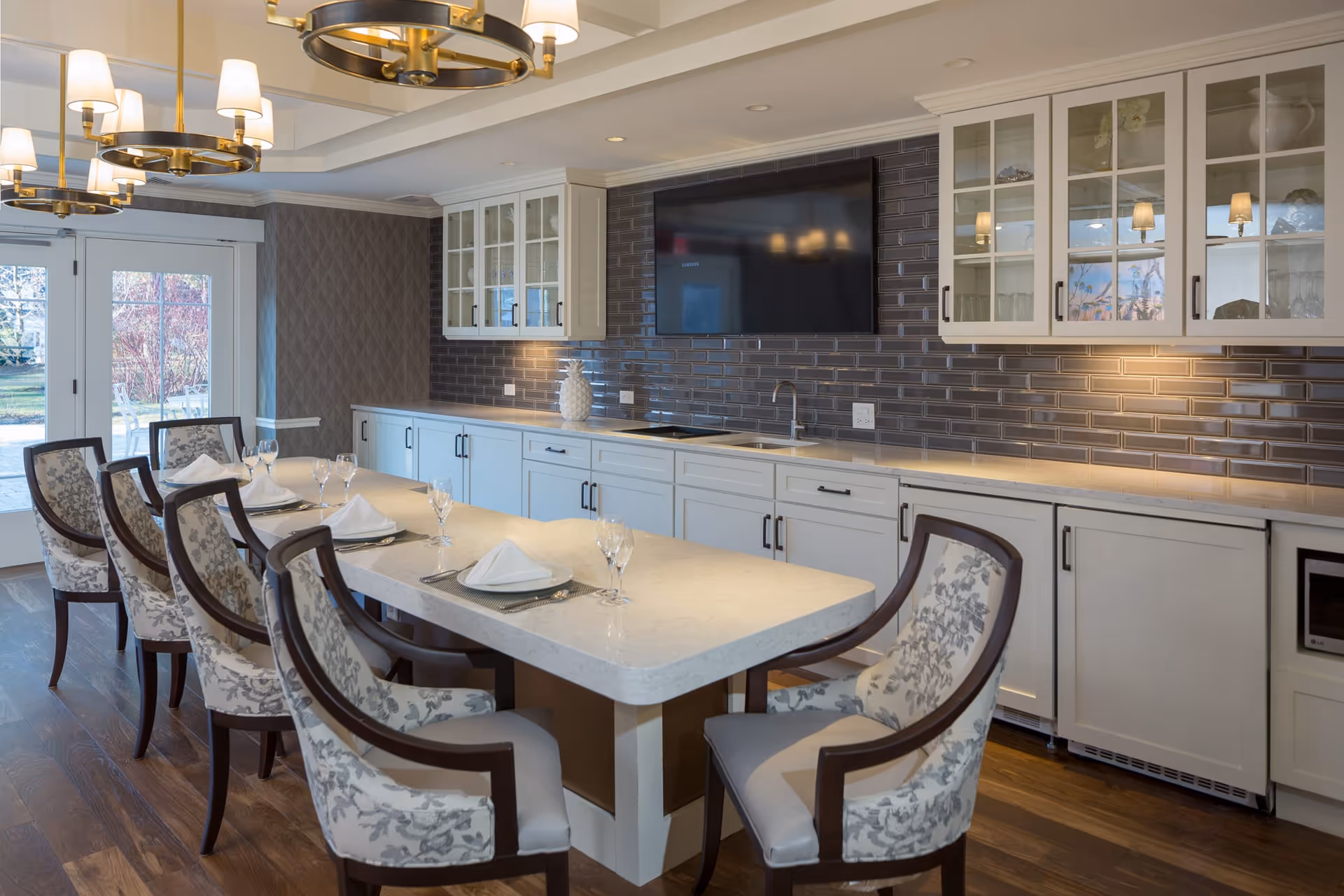 A modern dining area with a long white countertop table set with plates, napkins, and wine glasses. Six upholstered chairs with floral patterns surround the table. The background features white cabinetry with glass doors, a dark tiled backsplash, a sink, and a mounted flat-screen TV. Large windows and glass doors allow natural light to enter the room.