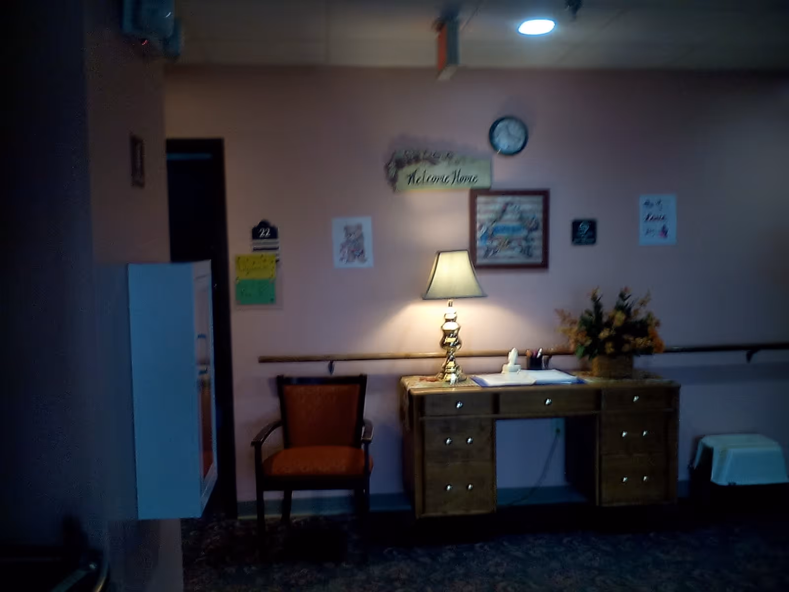 A dimly lit hallway area with a wooden desk holding a lamp, papers, and a flower arrangement. There is an orange upholstered chair next to the desk. On the wall above the desk are a clock, a framed picture, a 'Welcome Home' sign, and some other small decorations. The walls are painted light pink, and there is a handrail running along the wall.