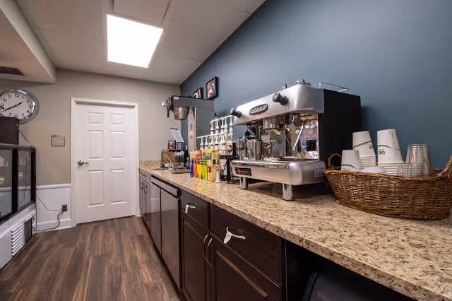 A coffee station area with a large espresso machine, syrup bottles, disposable cups in a wicker basket, and a coffee grinder on a granite countertop. The room has dark wood cabinets, a blue wall, a white door, and a clock on the wall.