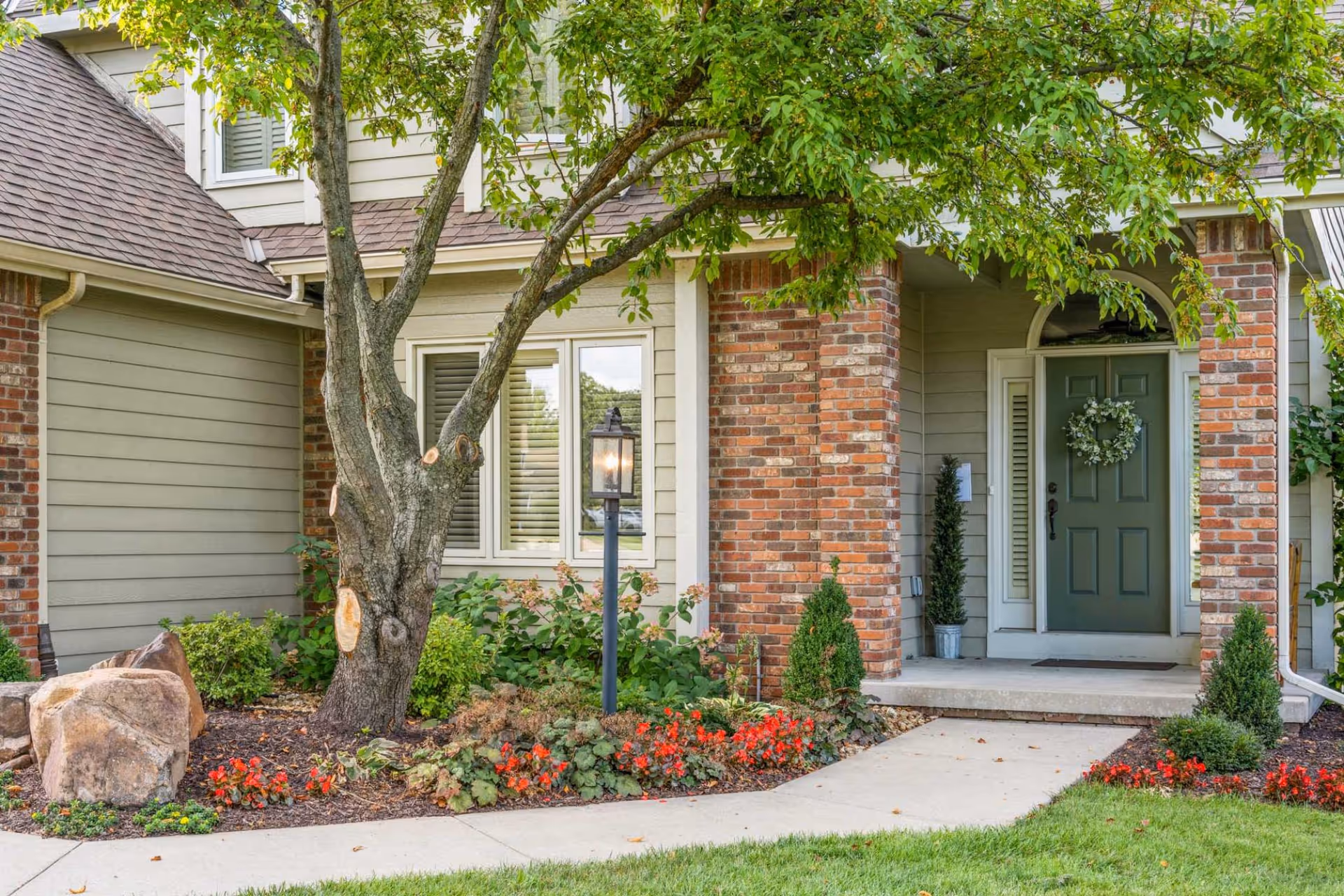 Front entrance of a brick and siding building with a green door, a tree, a lamp post, and landscaped flowerbeds along a concrete walkway.