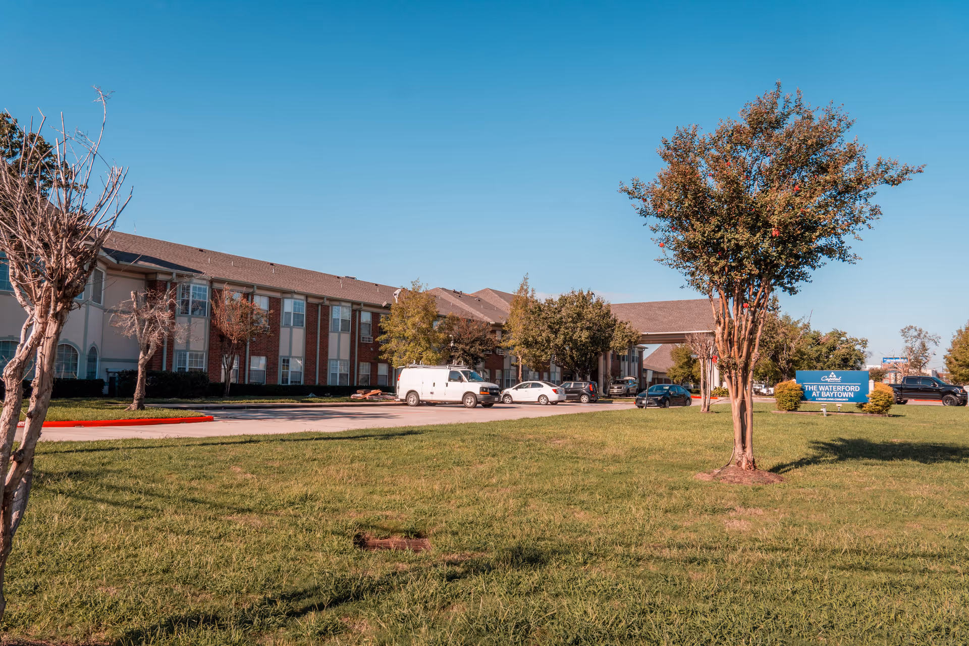 Exterior view of The Waterford at Baytown senior living facility showing a two-story building with a brick and beige facade, several parked cars, a green lawn with trees, and a blue sign with the facility's name under a clear blue sky.