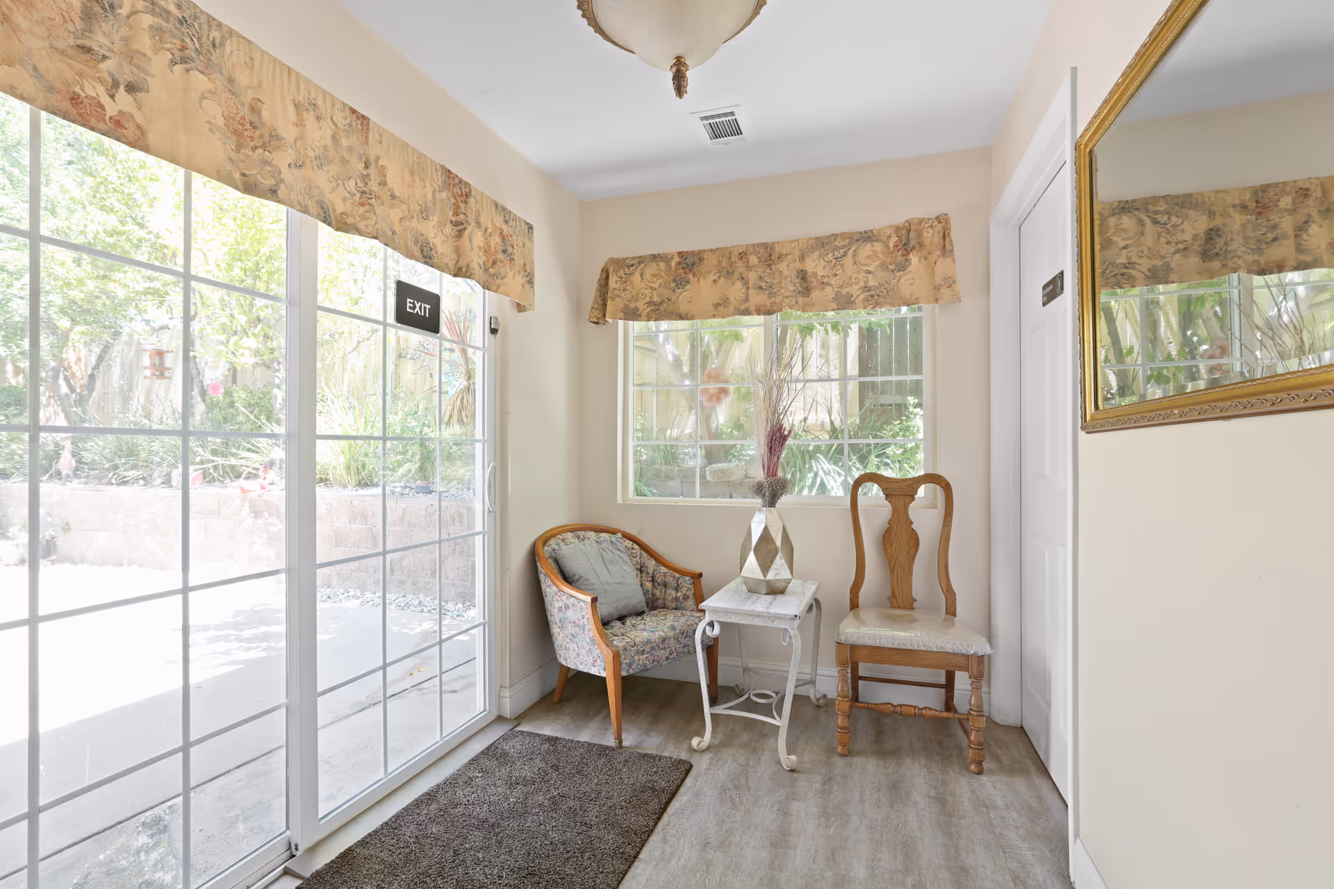 A small sitting area near a glass sliding door with an exit sign. The area has two chairs, one upholstered with floral fabric and a wooden chair with a cushioned seat, separated by a small white table with a decorative vase containing dried flowers. There is a window with floral valances matching those on the sliding door, and a large gold-framed mirror on the adjacent wall. Outside, greenery is visible through the glass door and window.