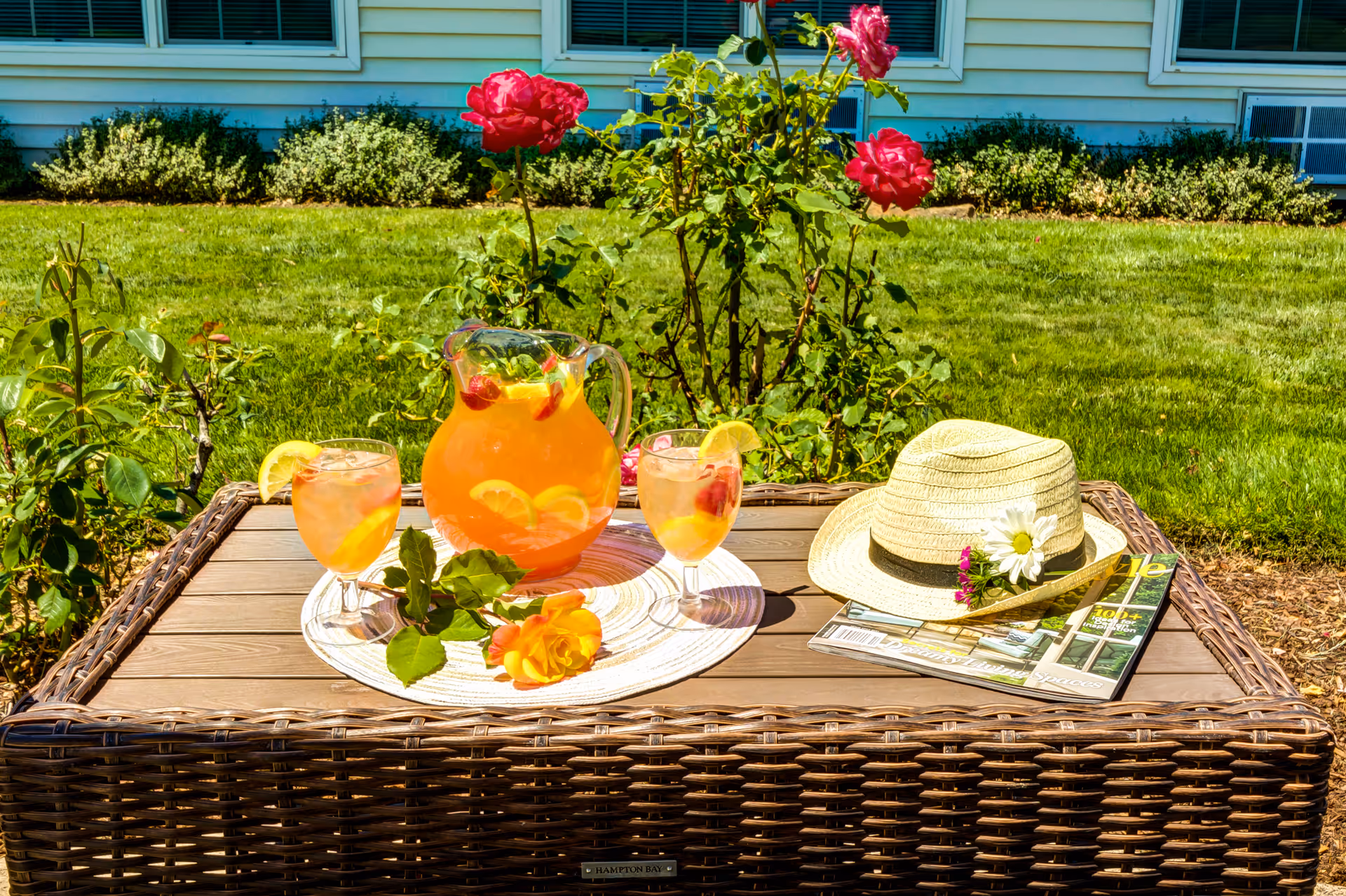 Outdoor wicker table with a pitcher of fruit-infused drink and two glasses garnished with lemon slices, a straw hat decorated with flowers, and magazines. The table is set in a garden with green grass, rose bushes with pink and yellow roses, and a building with windows in the background.