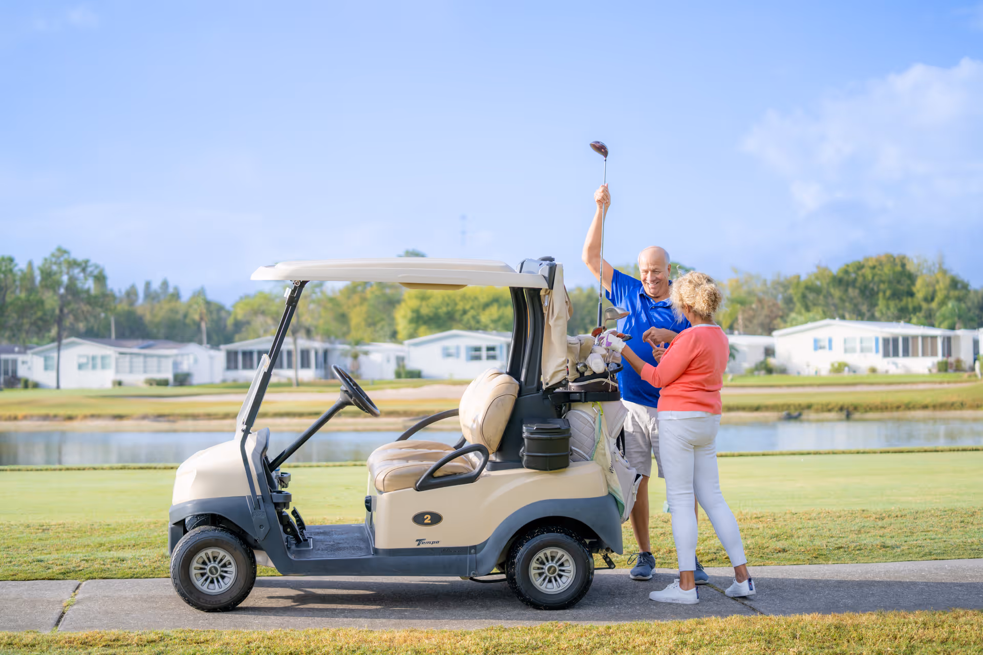 An elderly man and woman standing next to a golf cart on a golf course. The man is holding a golf club up in the air while the woman looks at the golf clubs in the cart. There are houses and trees in the background under a clear blue sky.