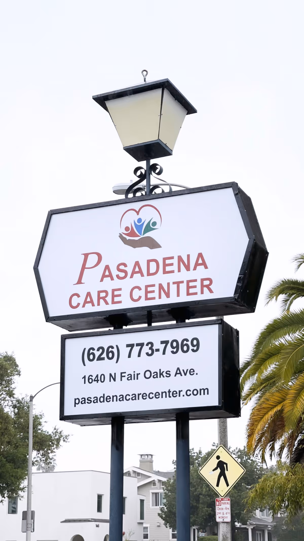 Outdoor sign for Pasadena Care Center with contact phone number, address, and website, mounted on a black post with a lamp on top. Trees and residential buildings are visible in the background.