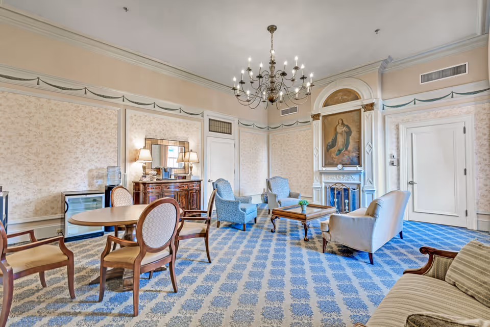 A spacious, elegantly decorated living room with patterned blue carpet and floral wallpaper. The room features a chandelier, a fireplace with a large painting above it, several upholstered chairs and sofas arranged around a wooden coffee table, and a round wooden table with chairs. There is a wooden sideboard with lamps and a mirror, and a water dispenser next to it.