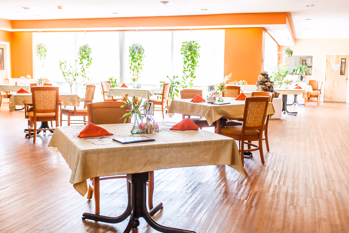 Bright dining room with multiple set tables, chairs, and plants near large windows.