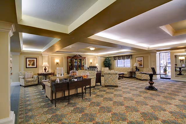 A spacious, well-lit common lounge with upholstered seating, tables, decorative cabinets, and patterned carpet.