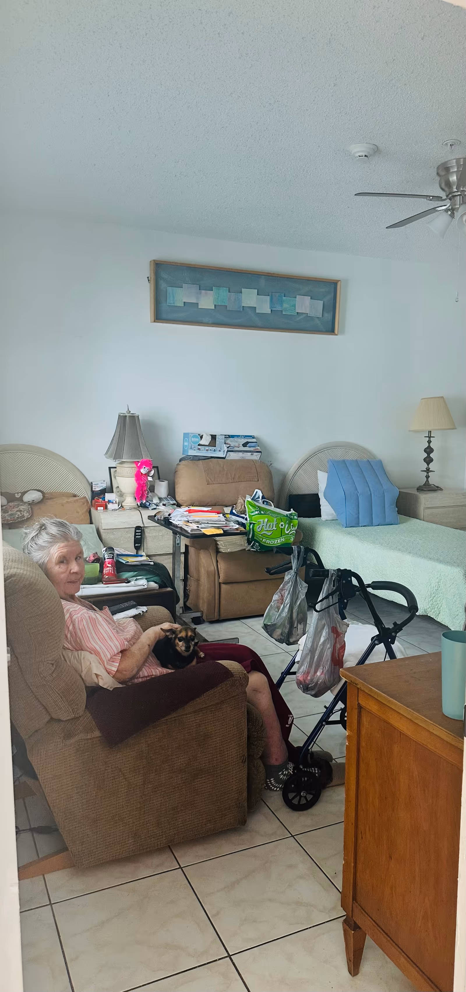 An elderly woman sitting in a brown recliner chair holding a small dog in a room with a bed, two nightstands with lamps, a walker with bags hanging on it, and various personal items on the tables. The room has tiled floors, a ceiling fan, and a framed abstract artwork on the wall.