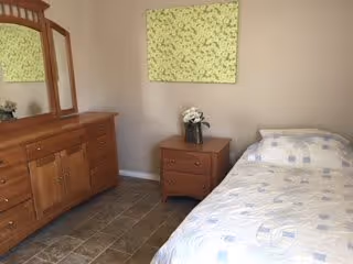 A simple bedroom with a single bed covered in a light-colored patterned bedspread, a wooden nightstand with a vase of white flowers, a wooden dresser with a mirror, and a green abstract painting on the wall.
