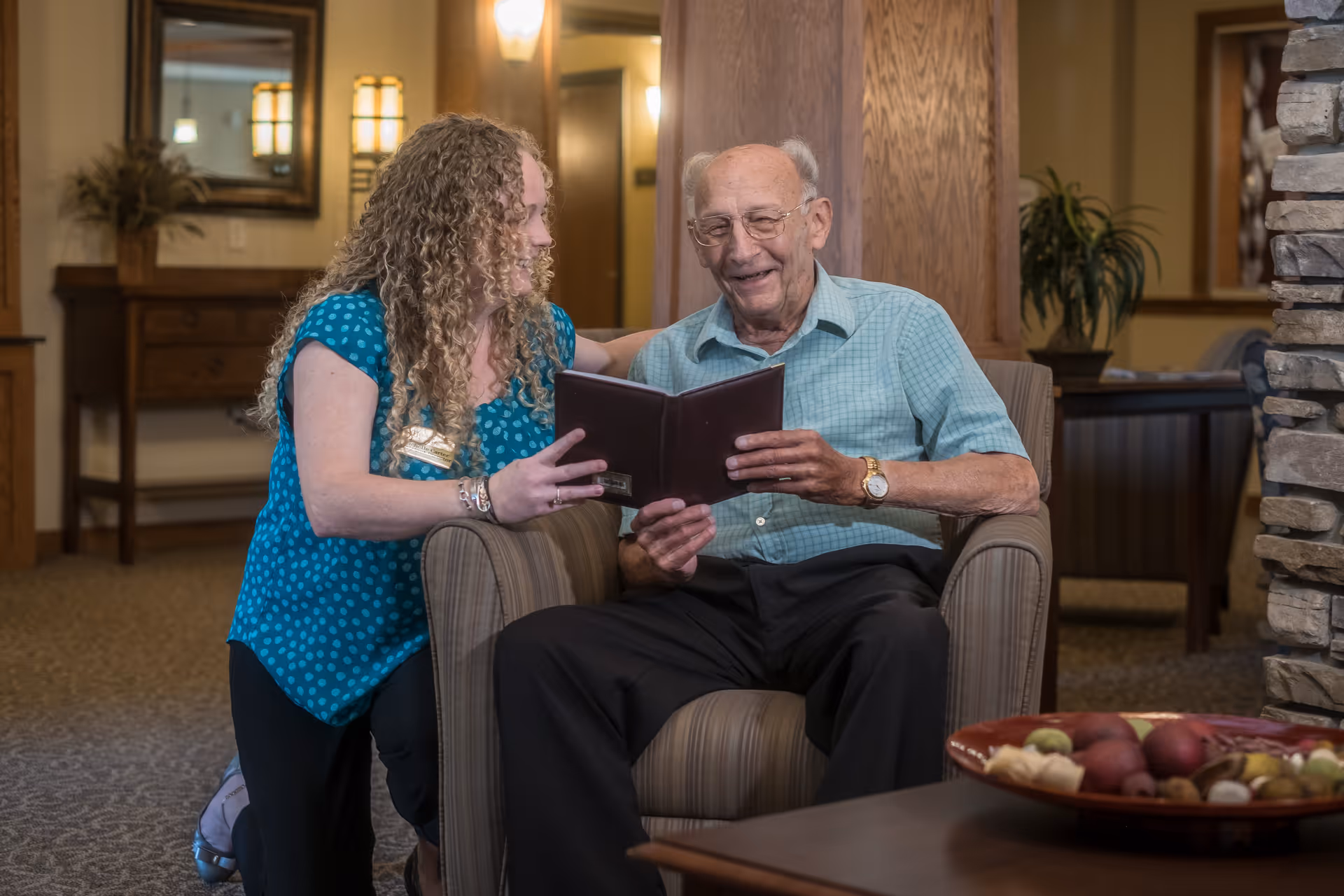 An elderly man sitting in a comfortable armchair reading a book with a smiling woman kneeling beside him in a warmly lit living room area with wooden furniture and a stone fireplace.