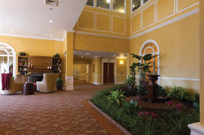Interior view of a senior living facility lobby area with yellow walls and patterned carpet. There is a seating area with two beige chairs and a small round table near a fireplace with shelves and plants. To the right, there is a raised indoor garden with various green plants and a small fountain. In the background, there is a hallway with elevators and framed pictures on the walls.