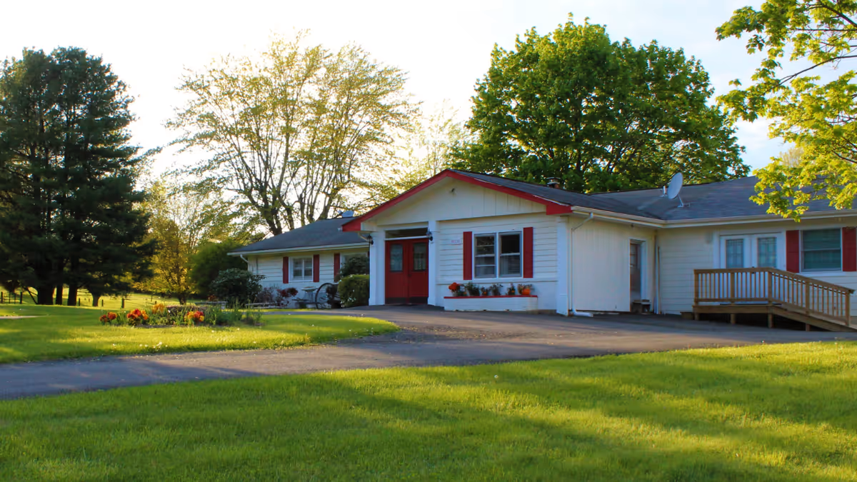 Single-story white assisted-living building with red trim and front doors, a driveway, ramp, and surrounding lawn and trees.