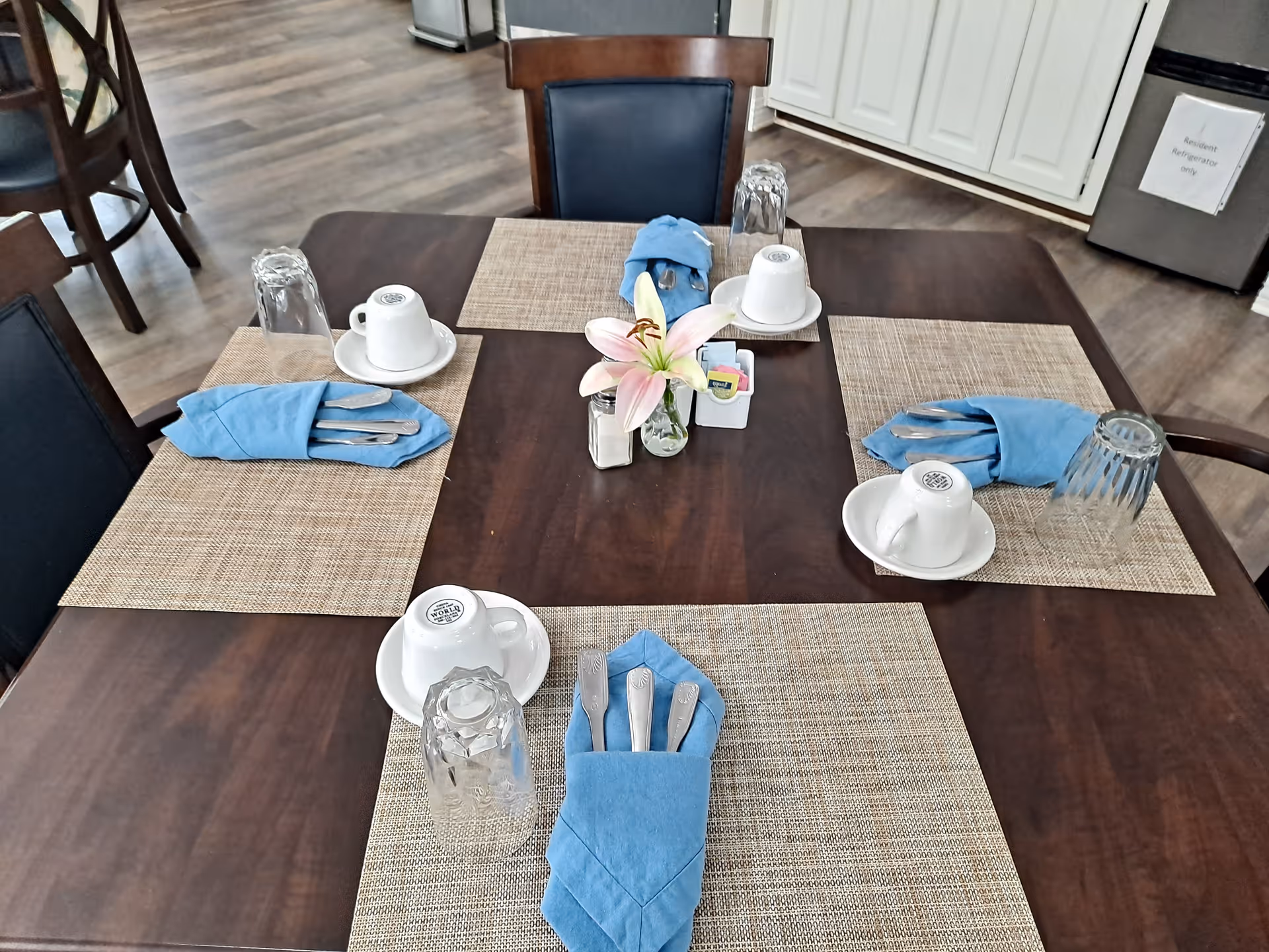 A dining table set for four with beige placemats, blue cloth napkins holding silverware, upside-down clear drinking glasses, white coffee cups on saucers, and a small vase with a pink lily flower in the center. The background shows wooden flooring, additional chairs, white cabinets, and a small refrigerator with a sign that reads 'Resident Refrigerator only.'