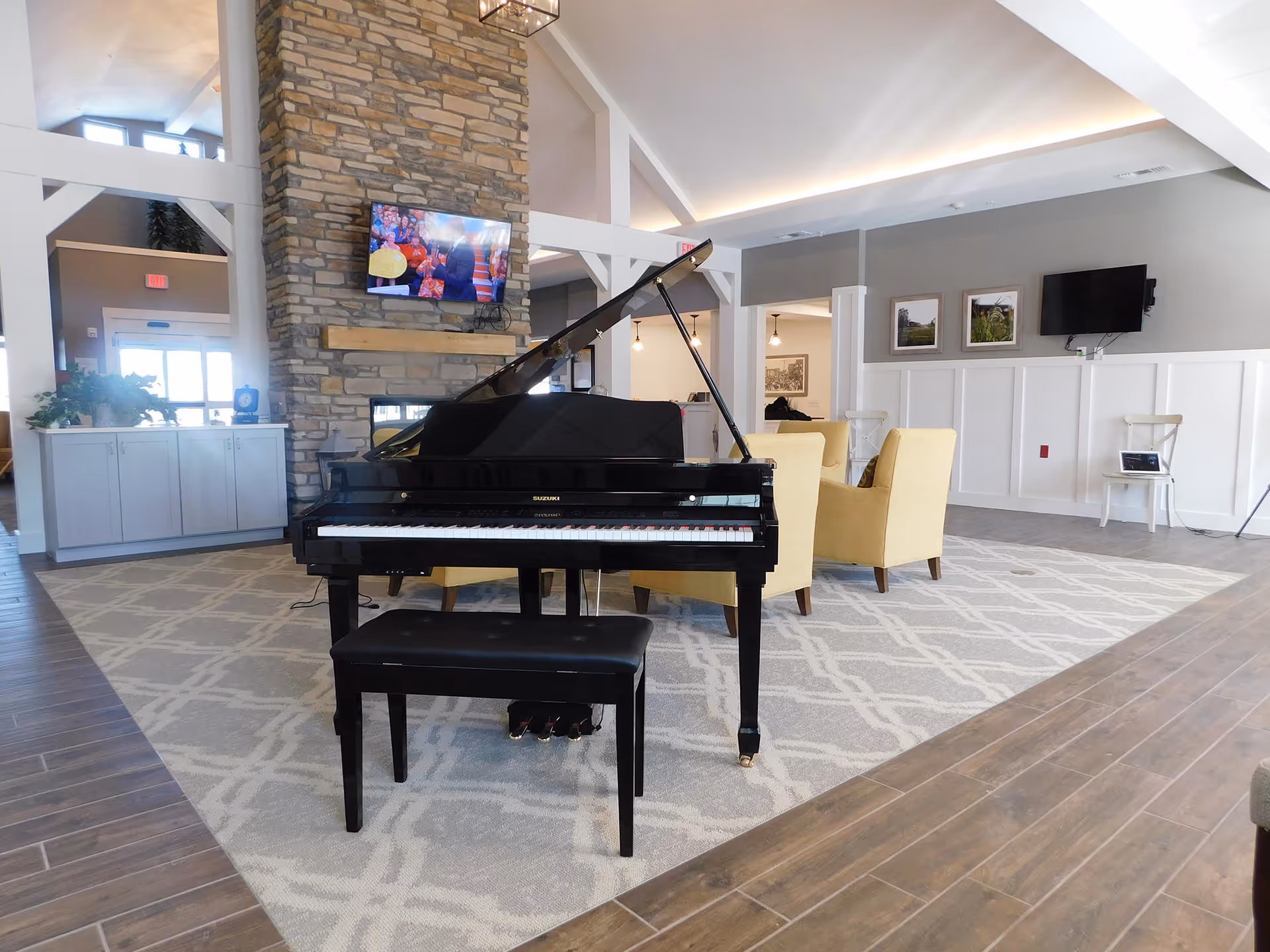 A spacious and well-lit living area featuring a black grand piano with a matching bench in the center on a patterned rug. Behind the piano, there is a stone fireplace with a mounted flat-screen TV showing a colorful scene. To the right, there are two yellow armchairs arranged for seating, and the walls are decorated with framed pictures and another wall-mounted TV. The room has wooden flooring and white wainscoting on the walls, with large windows allowing natural light to fill the space.