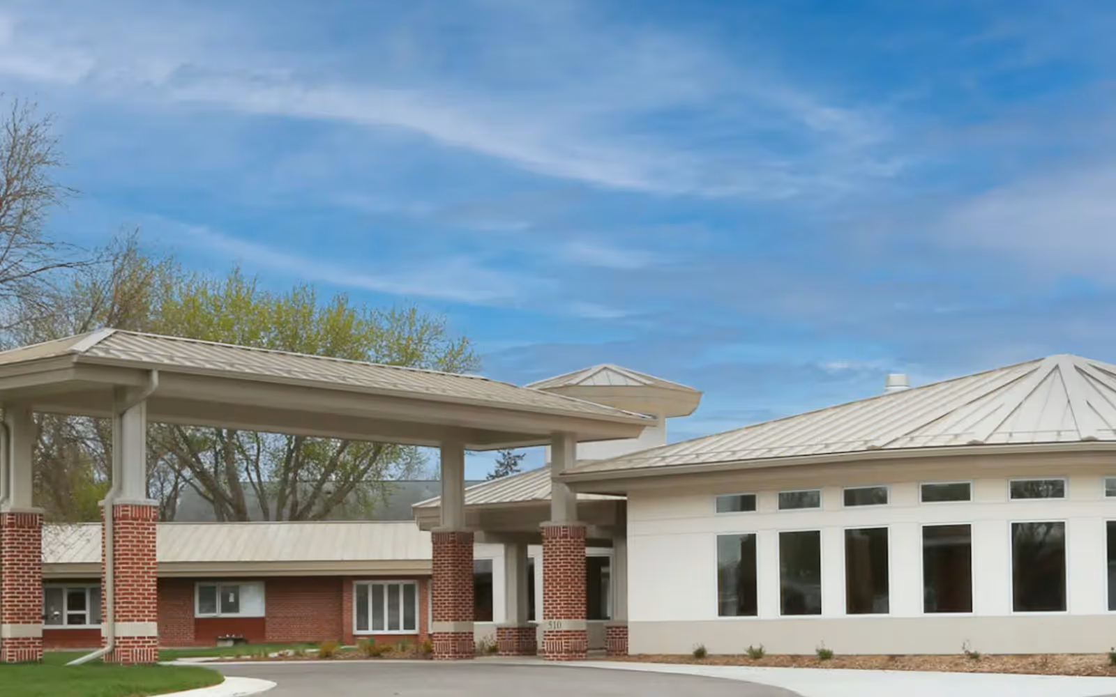 Front entrance of a single-story brick and stucco care building with a covered porte-cochere and rows of windows under a blue sky.