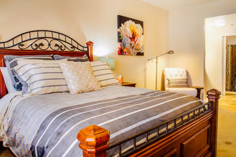 A well-appointed bedroom featuring a large wooden bed with striped bedding, pillows, a nightstand, armchair, lamp, and wall art.